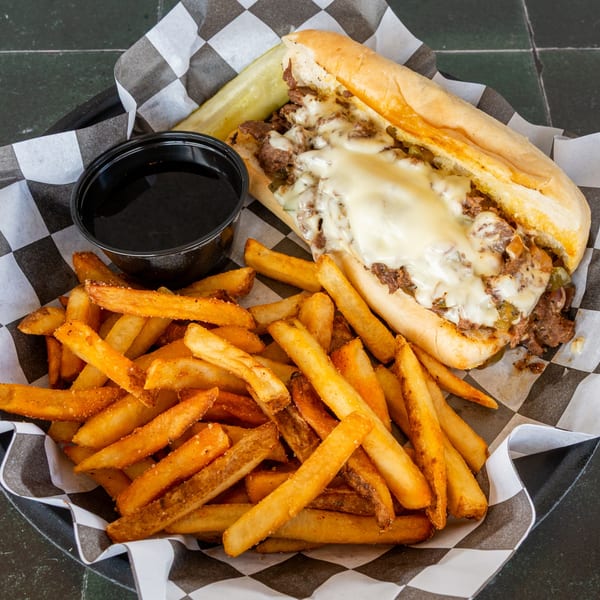 A cheesesteak sandwich with fries and a side of au jus in a checkered paper-lined basket.