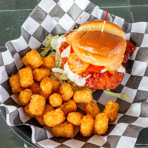 Spicy chicken sandwich with lettuce, tomato, pickles, and tater tots in a checkered basket.