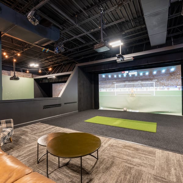 Indoor golf simulator bay with a large screen displaying a soccer game, seating, and coffee tables.