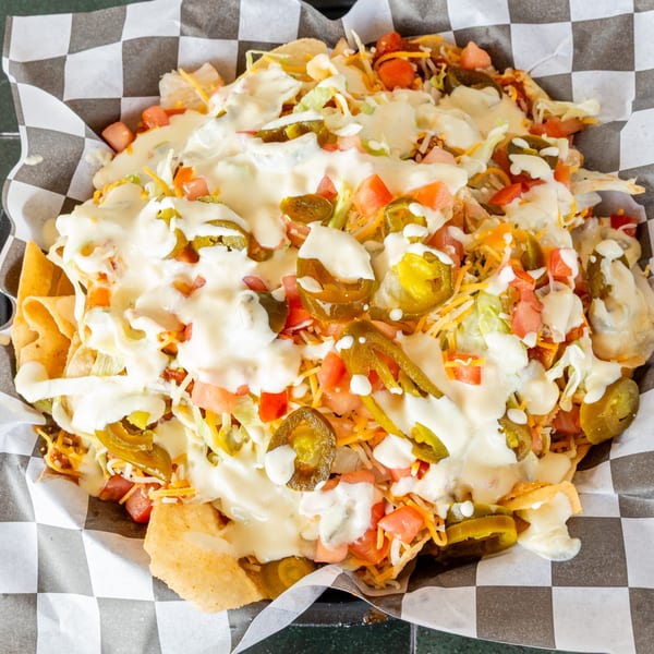 A large serving of nachos with melted cheese, jalapeños, tomatoes, and lettuce in a checkered paper-lined basket.