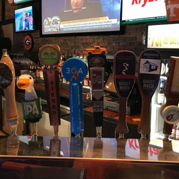 A row of diverse beer taps on a bar, with sports playing on TVs in the background.