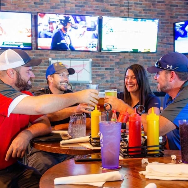 Four friends toasting beers at a table in 731 Sports Bar and Grill with TVs showing sports.